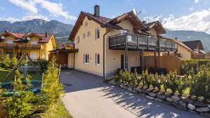 A two-story chalet with a balcony, a ski lift, and a mountain backdrop.