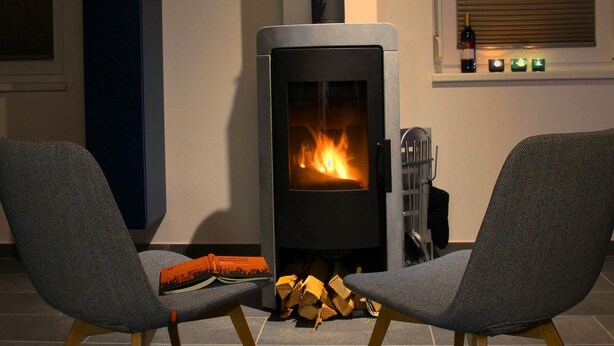 Un salon moderne avec un poêle à bois, deux fauteuils gris à revêtement en tissu, un livre et une bouteille de vin posés sur un rebord de fenêtre.