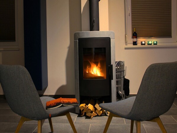 Un salón moderno con una estufa de leña, dos sillones grises tapizados, un libro y una botella de vino sobre un alféizar.