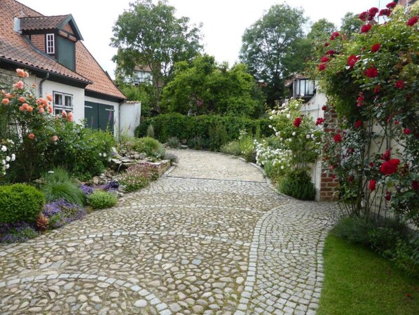 Un sentier pavé de pierres menant à travers un jardin où poussent différentes plantes à fleurs, et une maison en briques surmontée d’un toit en tuiles.