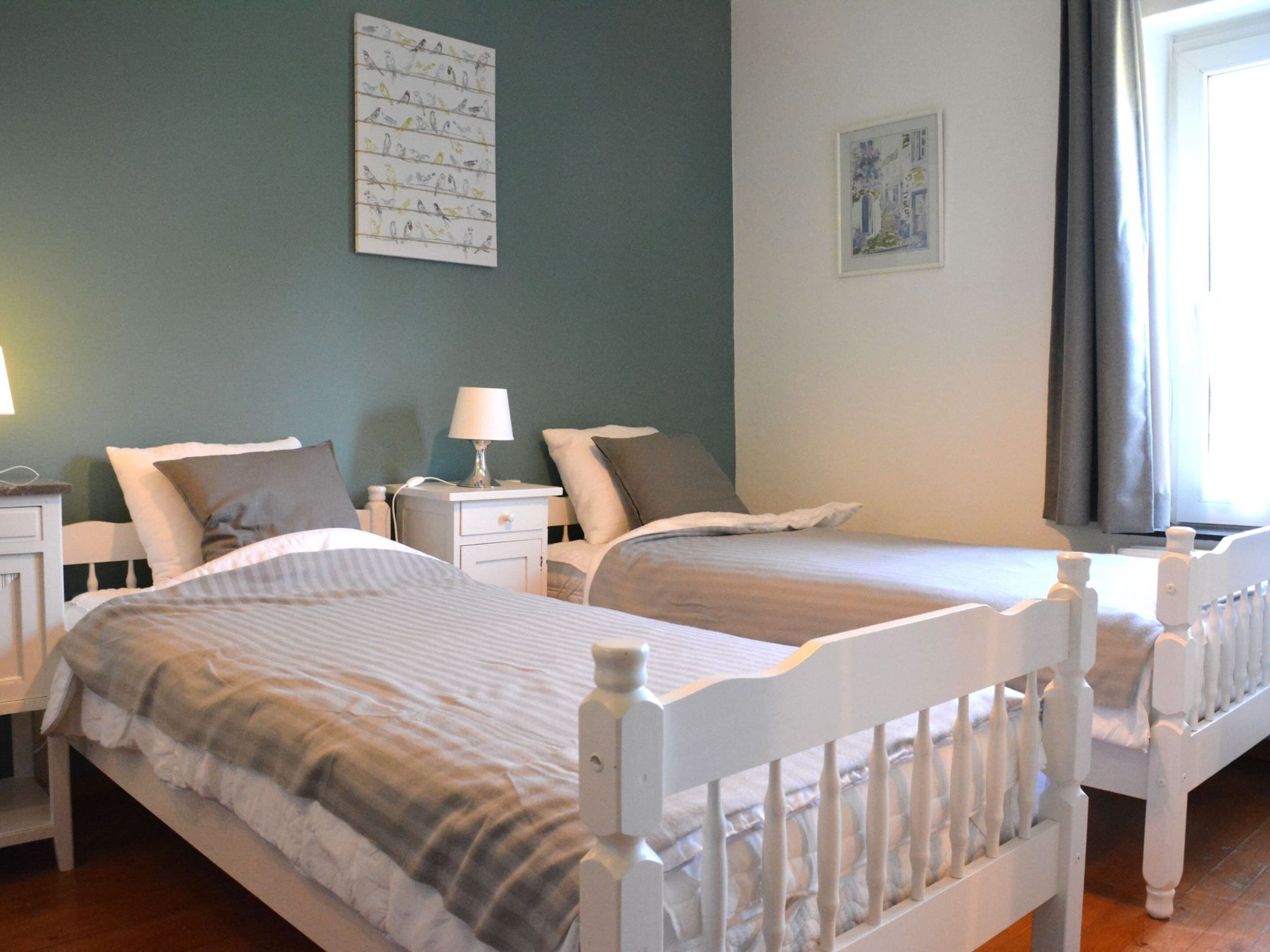 A hotel room with two single beds, a nightstand with a lamp, a wall-mounted artwork, and a window with curtains.