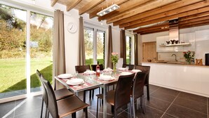 A dining area with a table set for four, a kitchen with white cabinets, and a view of a garden through large windows. - Cosy Holiday Home in Durbuy With Garden (Durbuy)