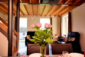 A dining area with a table set for two, a vase with pink roses, and a view of a living room with a sofa and a window.
