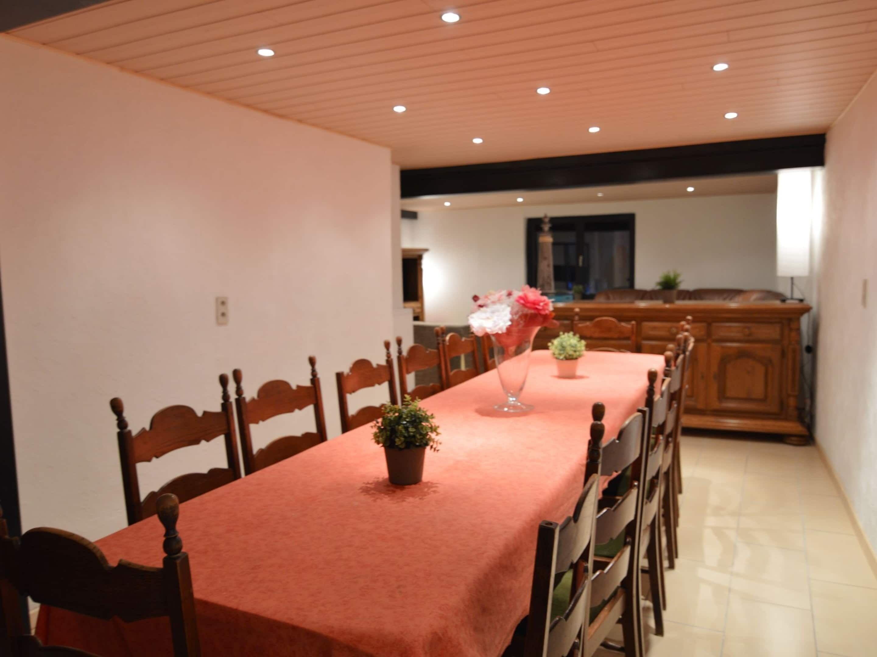 A dining area with a long table covered in a red tablecloth, wooden chairs, and potted plants.