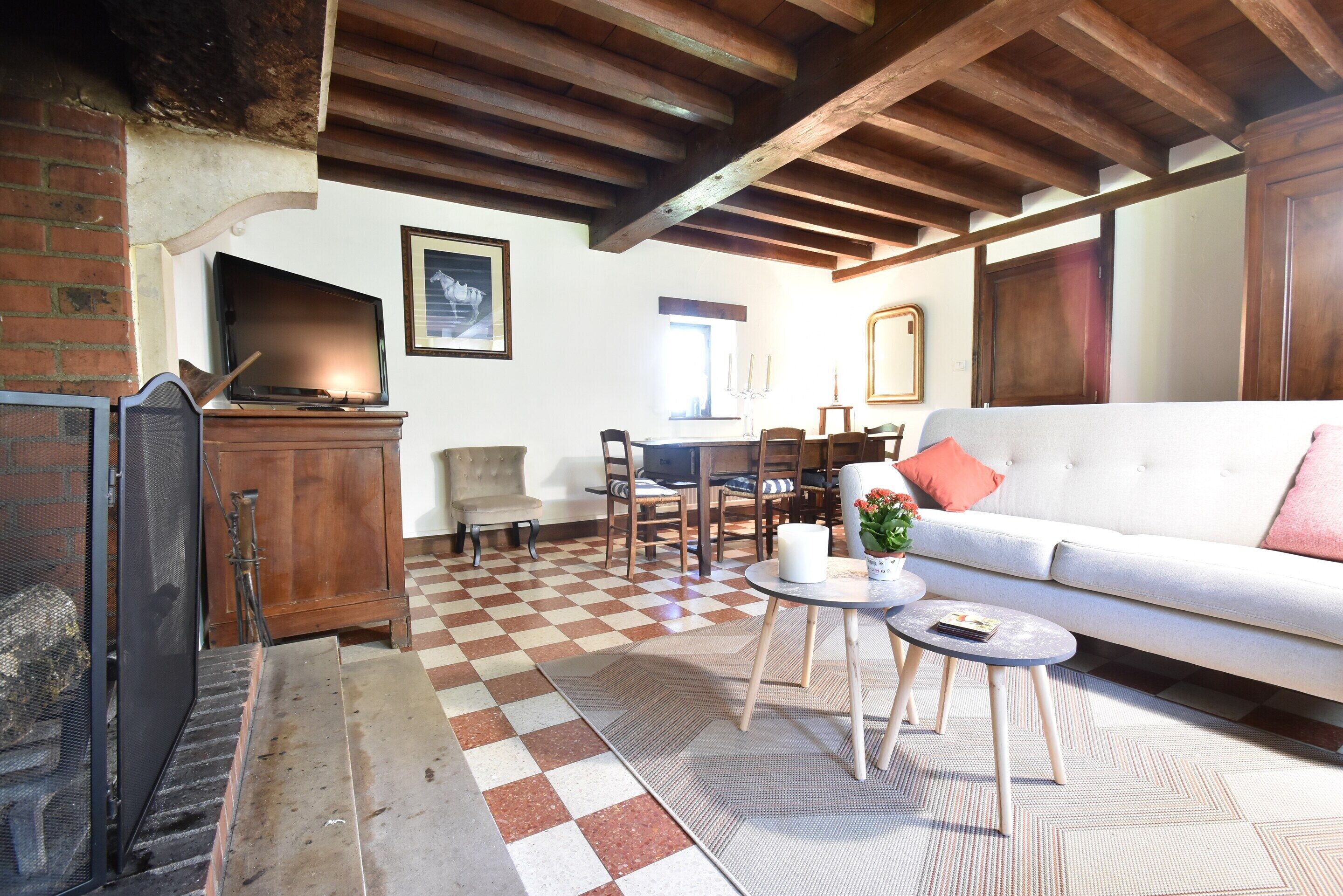 A living room with a fireplace, checkered floor, wooden beams, a sofa, a table, and a TV.