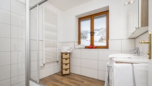 A modern bathroom with a shower, a window with a view of a snowy landscape, and a wooden shelf with towels. - Herzerl-Chalet Appartements (Flachau)