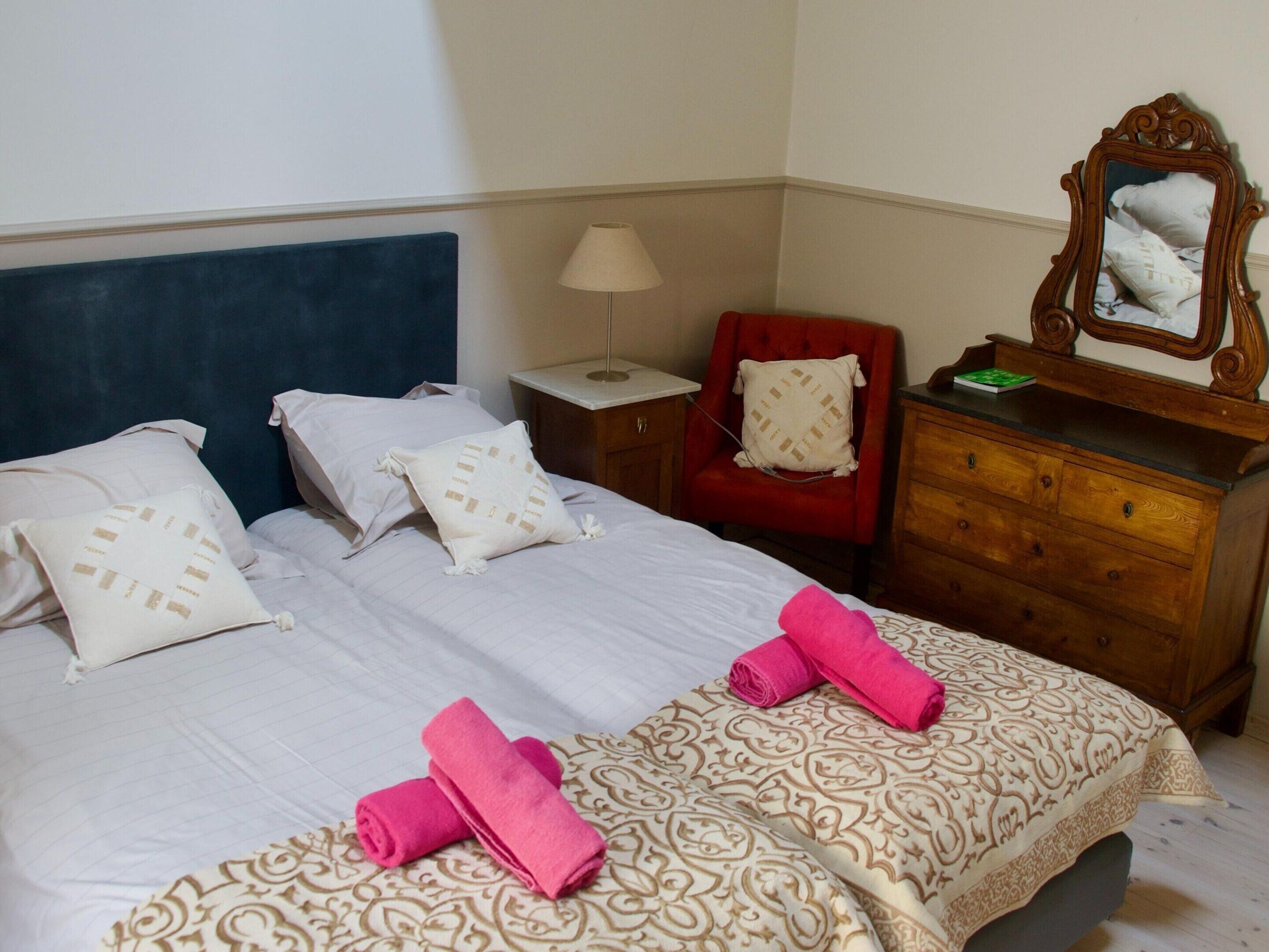 A neatly made bed with white linens and decorative pillows, a red armchair, a wooden dresser, and a mirror on the dresser.