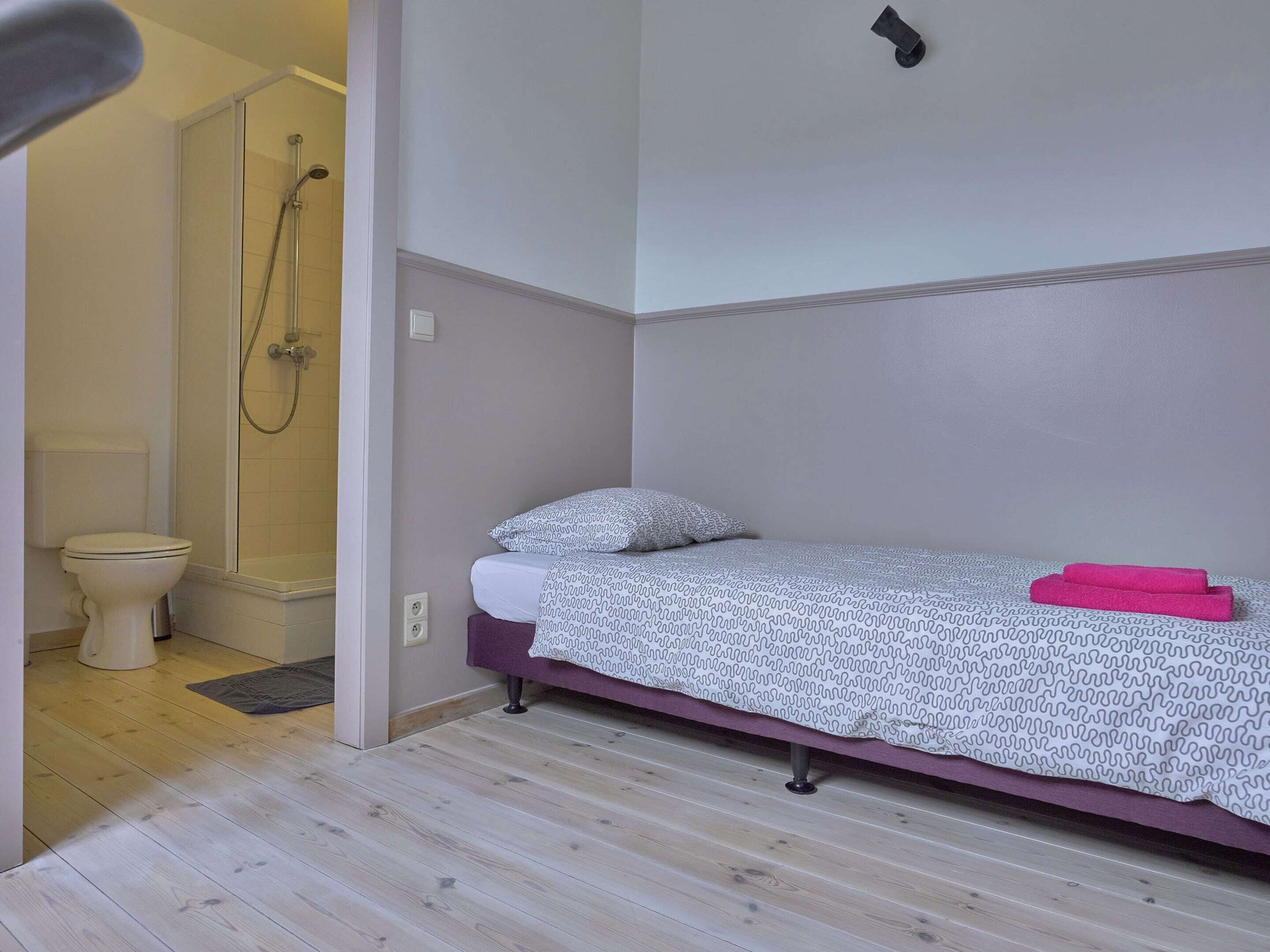 A small, single-bed room with a shower area, a toilet, and a purple towel on the bed.