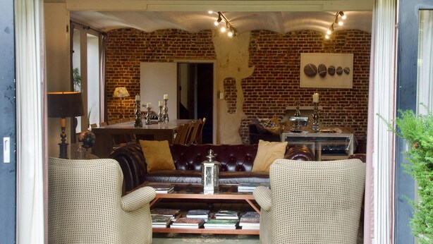 A cozy living room with a brick wall, a leather sofa, two wicker chairs, a coffee table, and a bookshelf. - House in Former Monastery (Sint-Truiden)