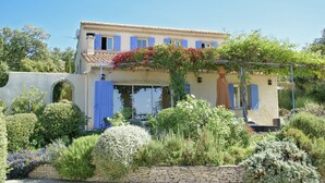Ein Gebäude im mediterranen Stil mit einem Ziegeldach, blauen Fensterläden und einem Garten mit verschiedenen Pflanzen und Blumen.