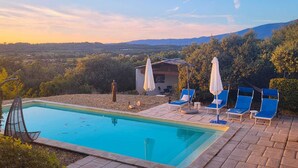 Ein Swimmingpool mit Liegestühlen und Sonnenschirmen, umgeben von Bäumen und Blick auf ferne Hügel.