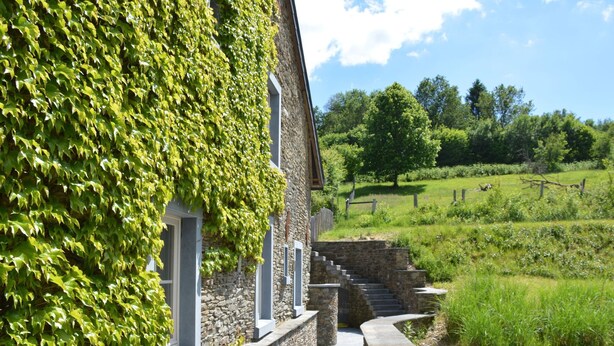 Un bâtiment en pierre avec un escalier en pierre menant à une porte, le tout entouré de verdure et sous un ciel dégagé.