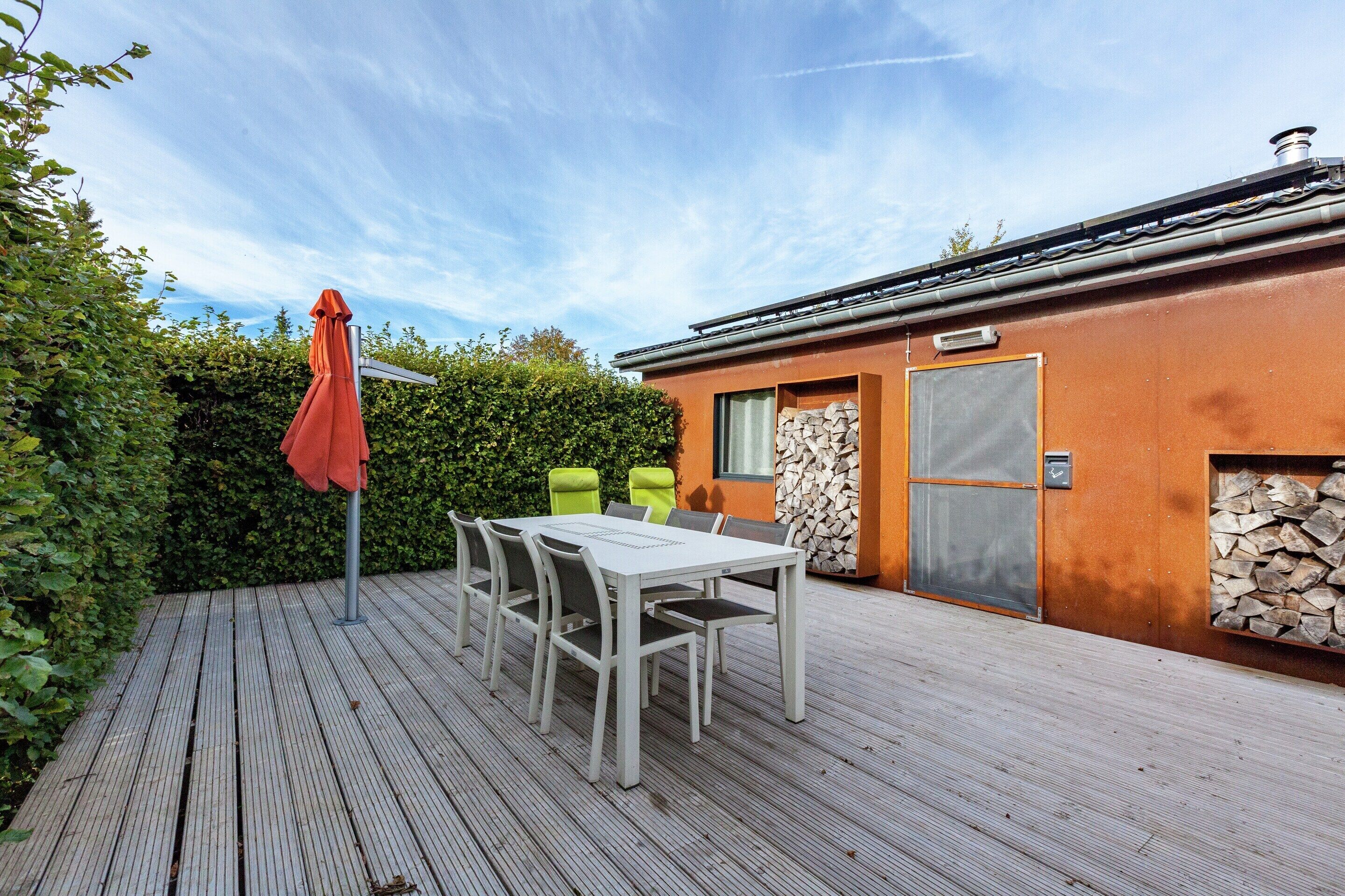A wooden deck with a table and chairs, a red umbrella, and a building with a stone wall and a door.
