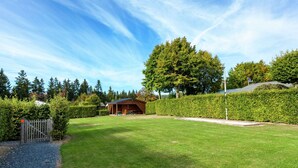 Une pelouse bien entretenue, agrémentée d’une structure en bois, probablement un gazebo ou un pavillon, et entourée de haies et d’arbres taillés avec soin.