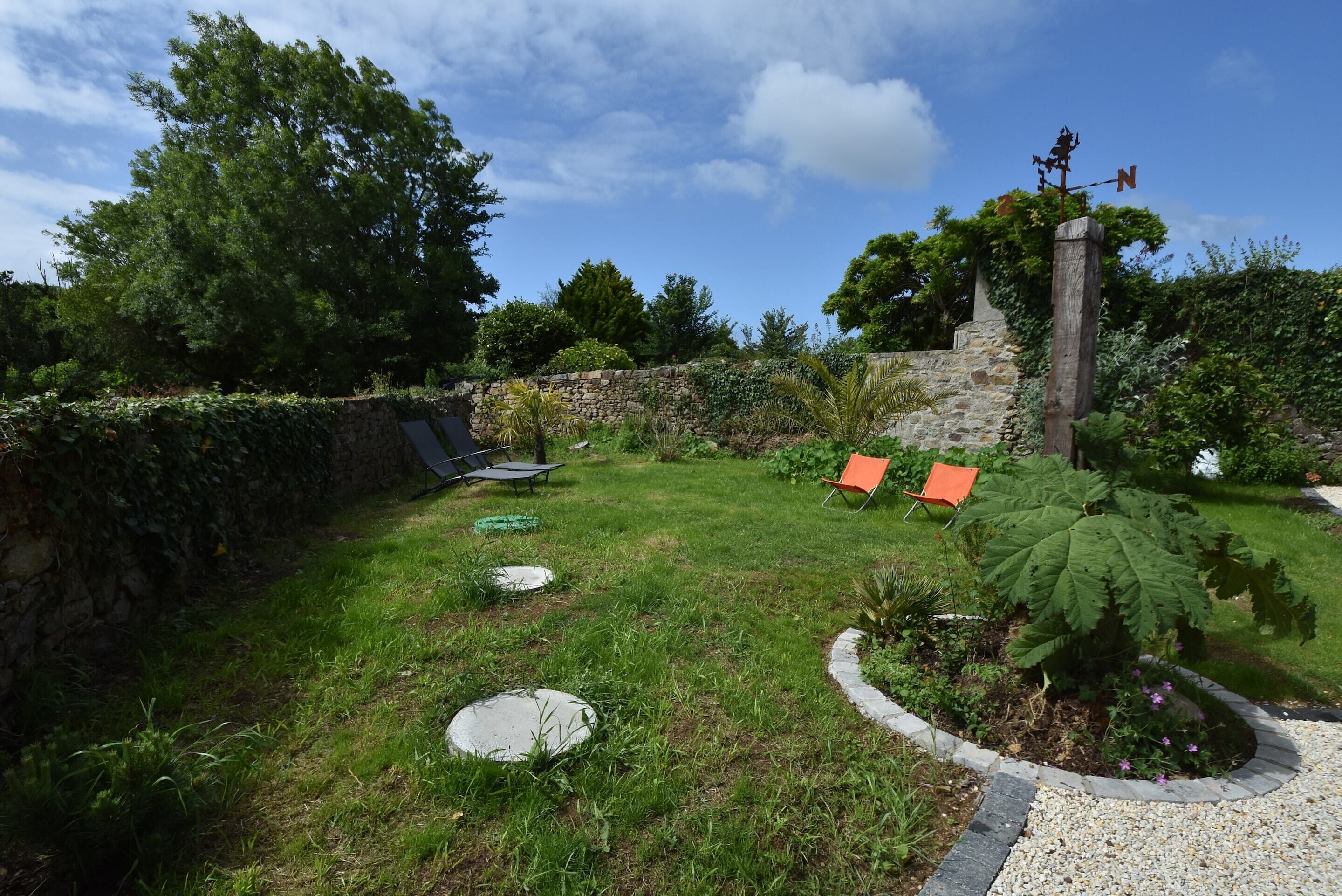 A garden with a stone wall, a weather vane, and a small tree.