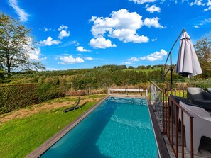 A swimming pool with a wooden deck and lounge chairs, surrounded by a hedge and a view of a forested hill. - Lush Chalet Near Lake of Butgenbach (Buellingen)