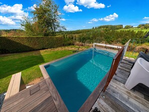 Ein rechteckiger Swimmingpool mit Holzterrasse und Metallgeländer, umgeben von einem gepflegten Rasen und einem Heckenstreifen.