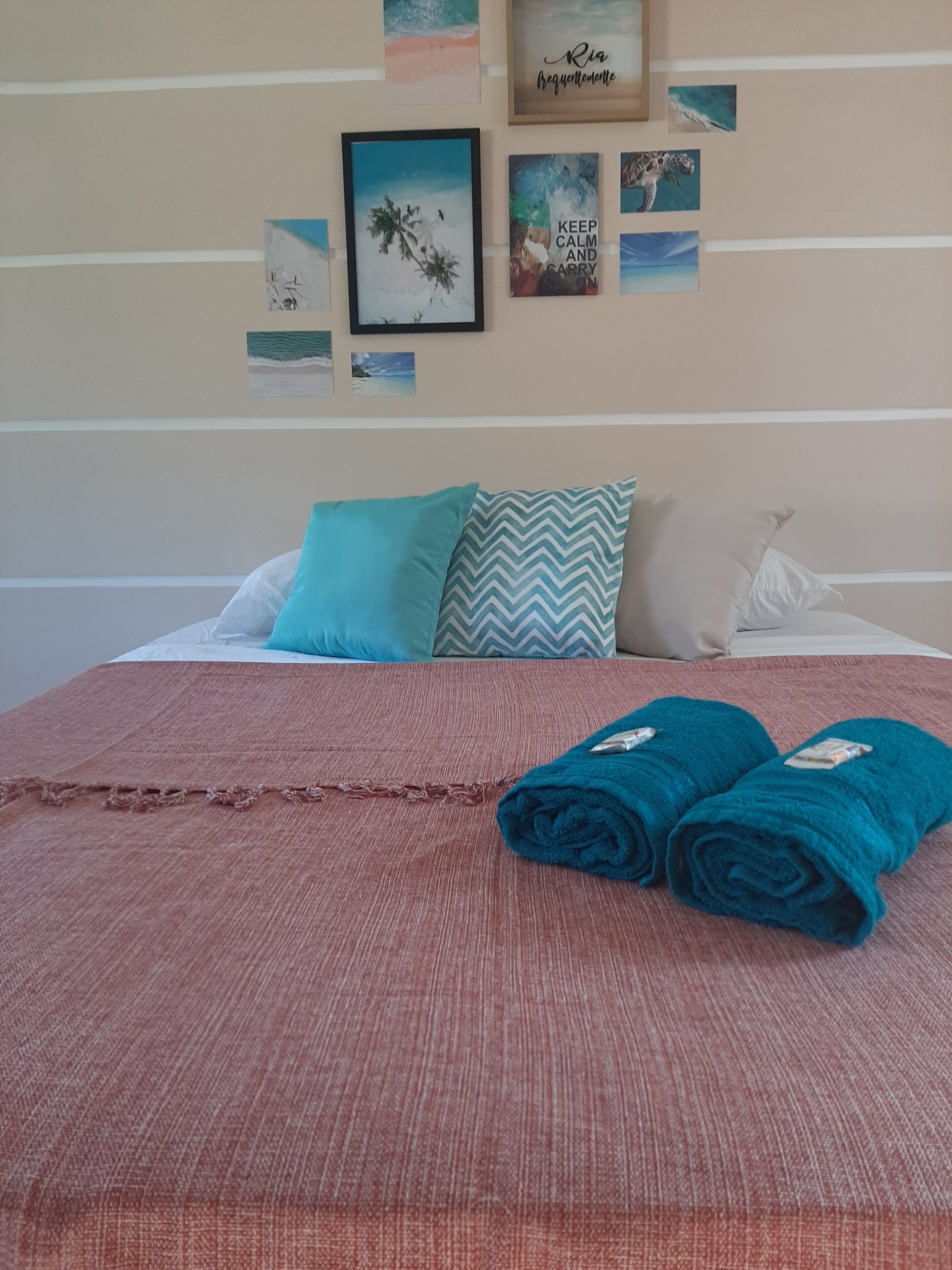 A neatly made bed with a red blanket and two rolled towels, a teal pillow, and a chevron-patterned pillow.
