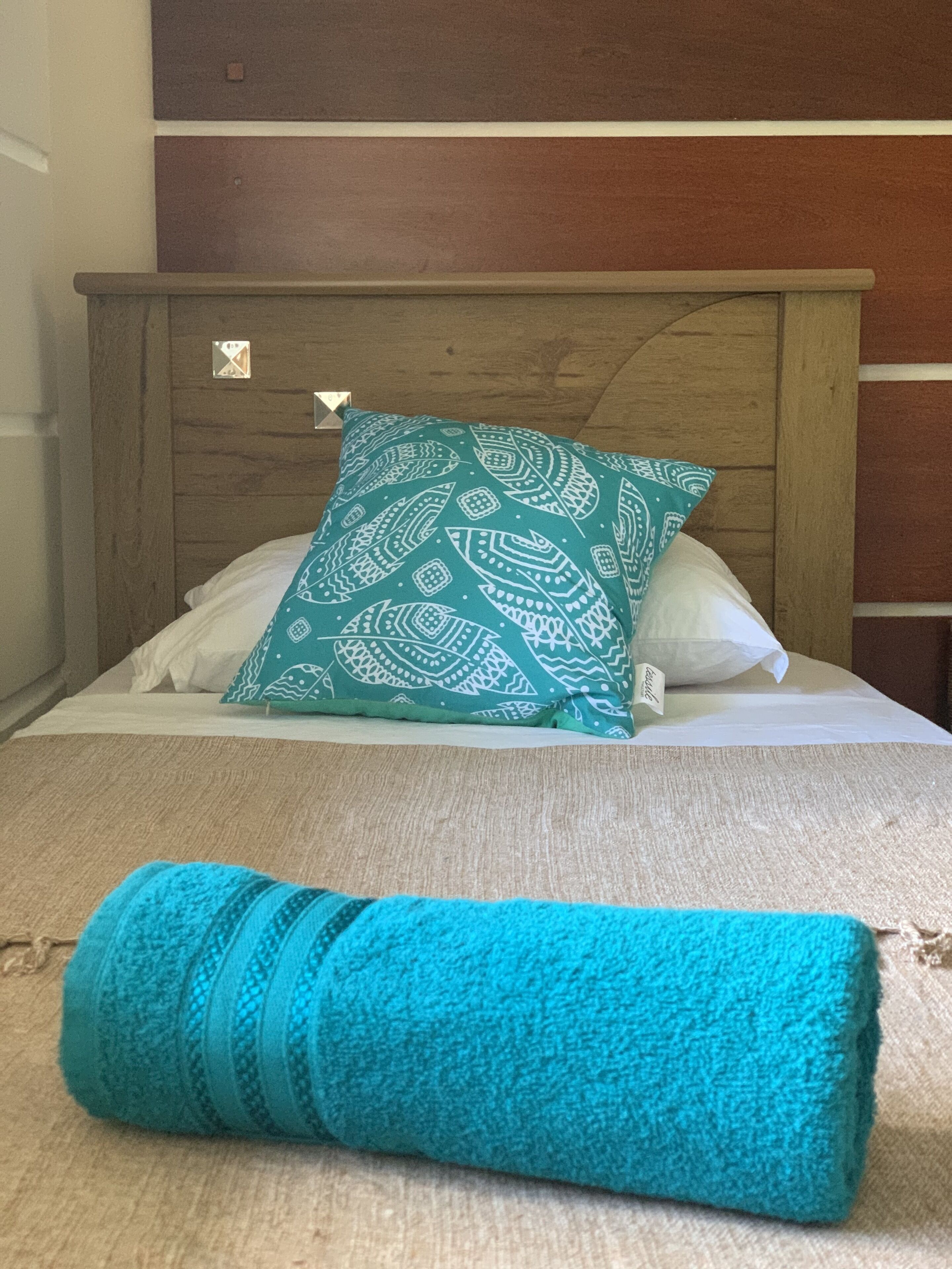 A bed with a wooden headboard, white pillows, and a teal patterned pillow. A rolled-up teal towel is on the bed.