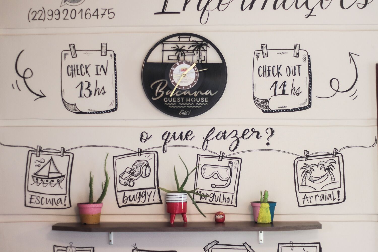 Una pared con un reloj que indica 'Bebana Guest House' y un letrero que dice 'Registro de entrada 13h, Registro de salida 11h'. Abajo, hay cuatro fotografías enmarcadas con las palabras 'Escura!', 'buggy!', 'Mergulho!', y 'Arraial!'.