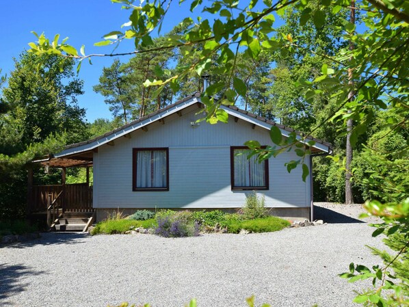 A single-story cabin with a gravel driveway, surrounded by trees and a wooden deck.