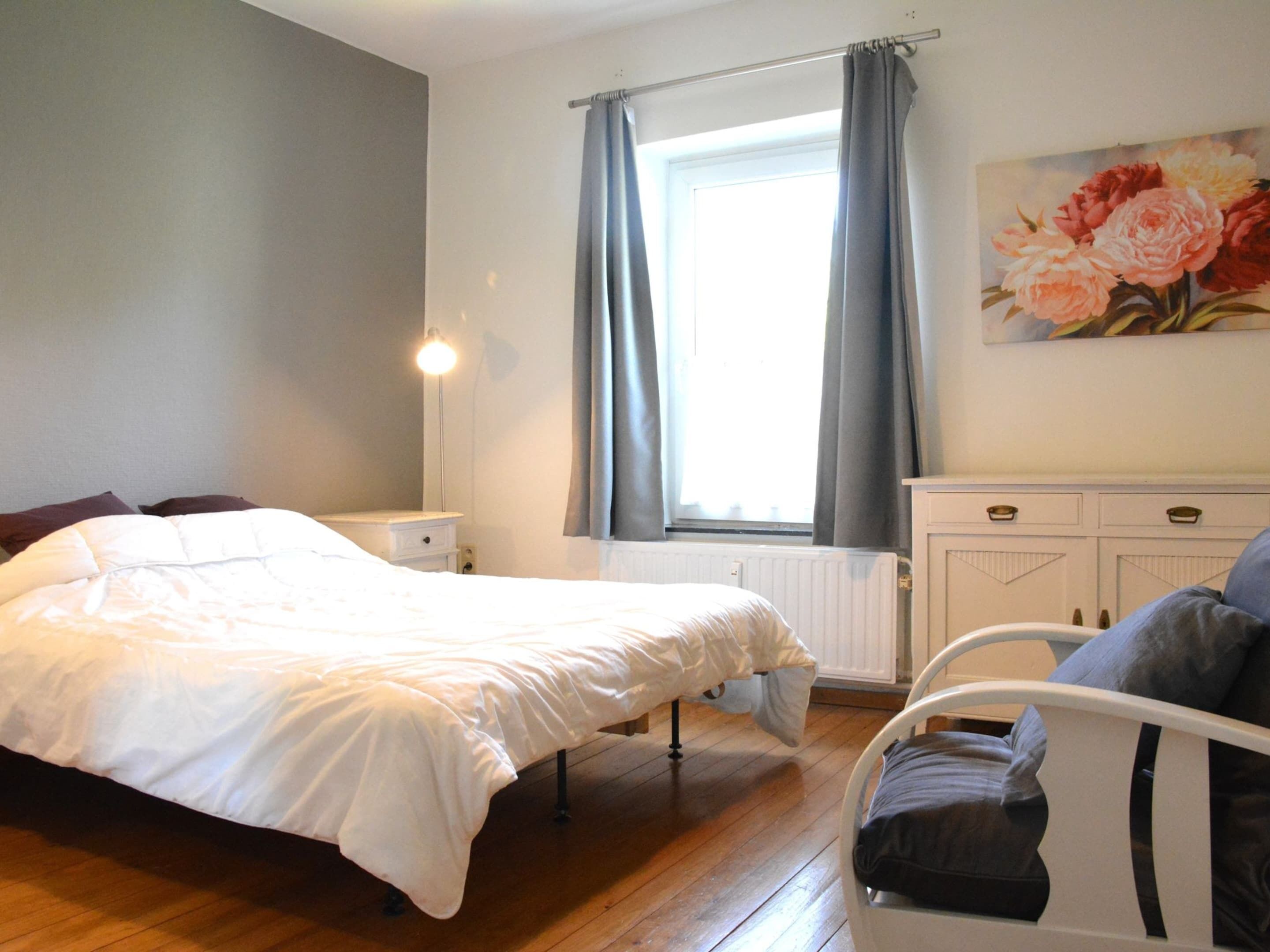 A bedroom with a bed, bedside table, nightstand, radiator, window with curtains, and a painting on the wall.