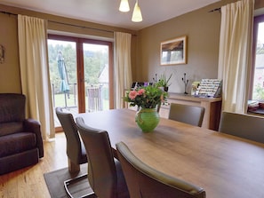 Un coin repas avec une table en bois, des chaises, un vase vert rempli de fleurs, un tableau encadré au mur et une vue à travers la porte vitrée.
