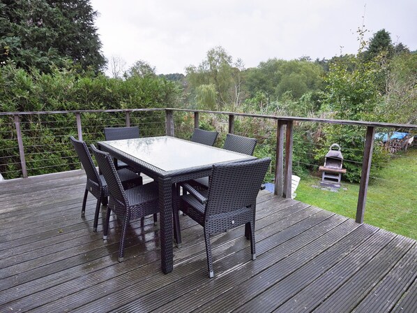 Une terrasse avec une table et des chaises, entourée d’une rambarde en bois et de verdure.