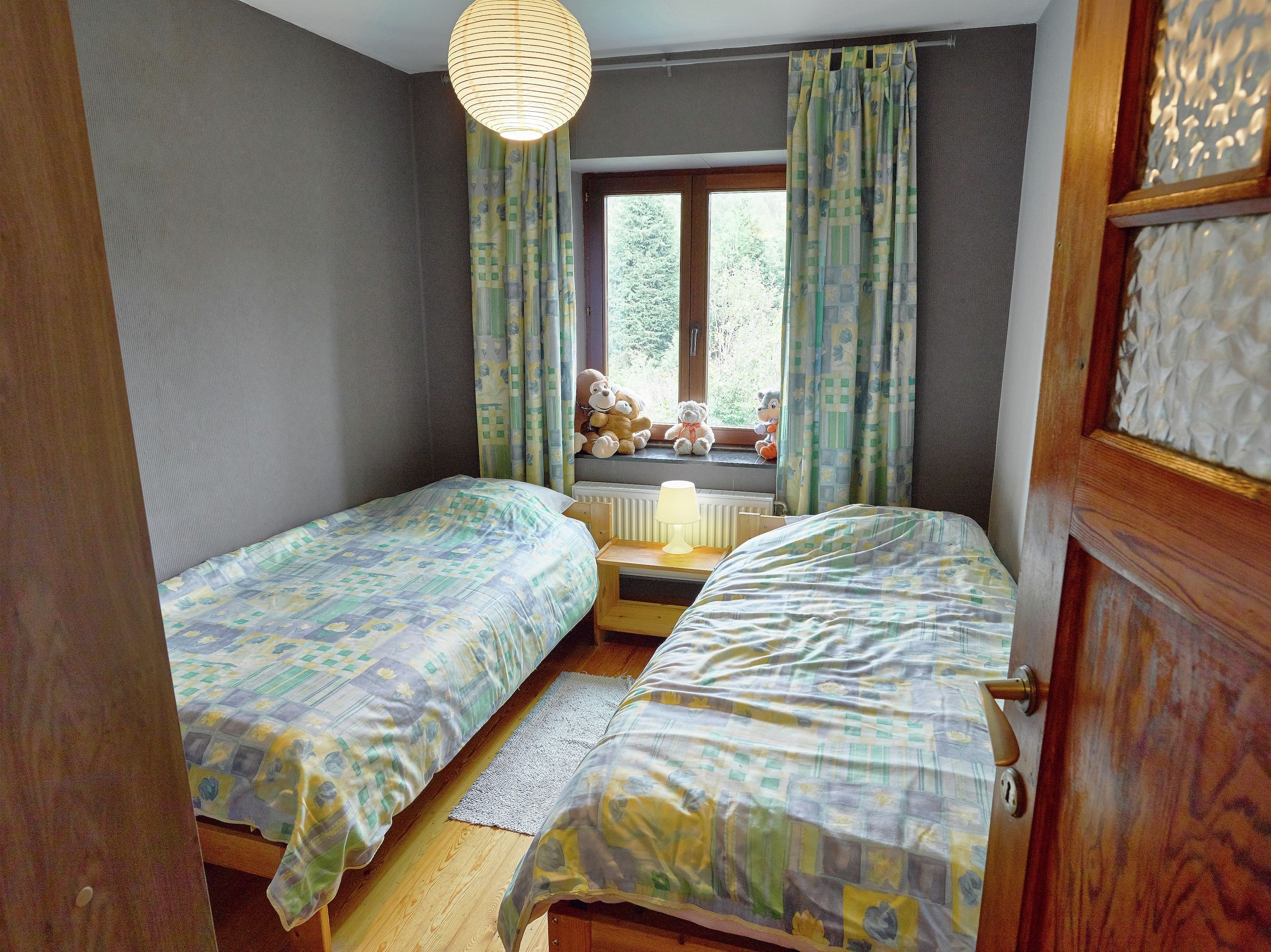 Une chambre avec deux lits, une fenêtre avec des rideaux, une lampe sur une table de chevet et des peluches sur le rebord de la fenêtre.