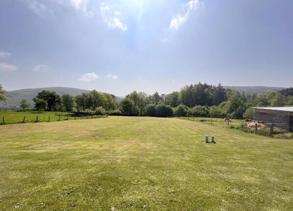 Ein grasbewachsenes Feld mit einem eingezäunten Bereich, einem kleinen Gebäude und Bäumen im Hintergrund.