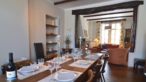 Uma sala de jantar com uma mesa de madeira posta para seis pessoas, rodeada por cadeiras. Há uma garrafa de vinho Merlot sobre a mesa. O quarto possui vigas de madeira no teto e uma janela com cortinas.
