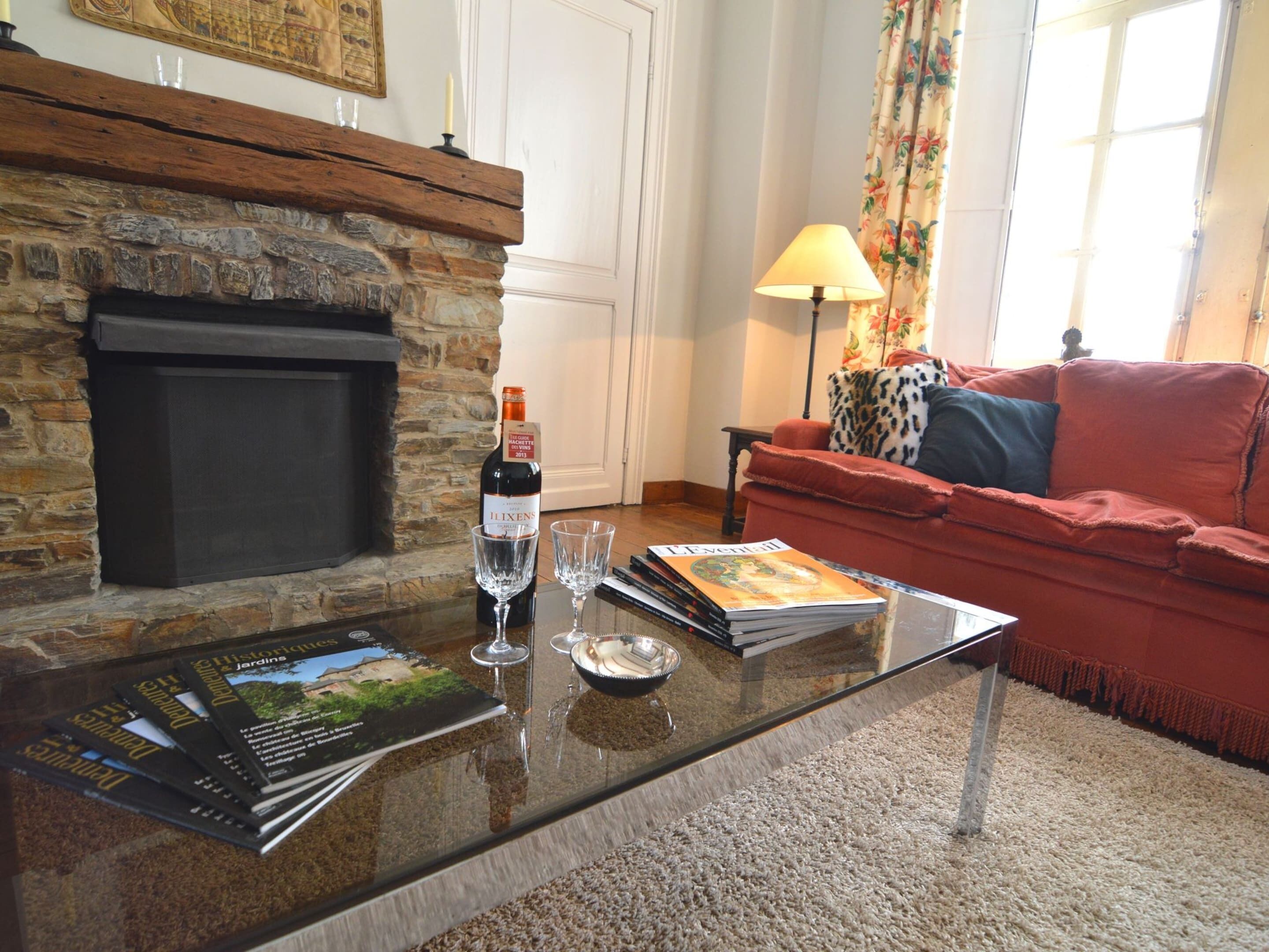 Un salón con chimenea de piedra, un sofá rojo, una mesa de centro de cristal con copas de vino y revistas, y una lámpara.