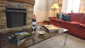 Uma sala de estar com lareira de pedra, um sofá vermelho, uma mesa de centro de vidro com copos de vinho e revistas, e um abajur.