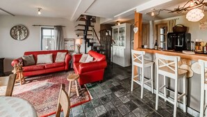 Uma sala de estar acolhedora com um sofá vermelho, uma pequena mesa de madeira e um tapete estampado. Há uma área de cozinha com um balcão e cadeiras brancas.