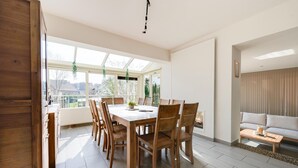 Een ruime eethoek met een grote tafel en stoelen, een oranjerie met glazen wanden en een woonkamer met een bank en kussens.