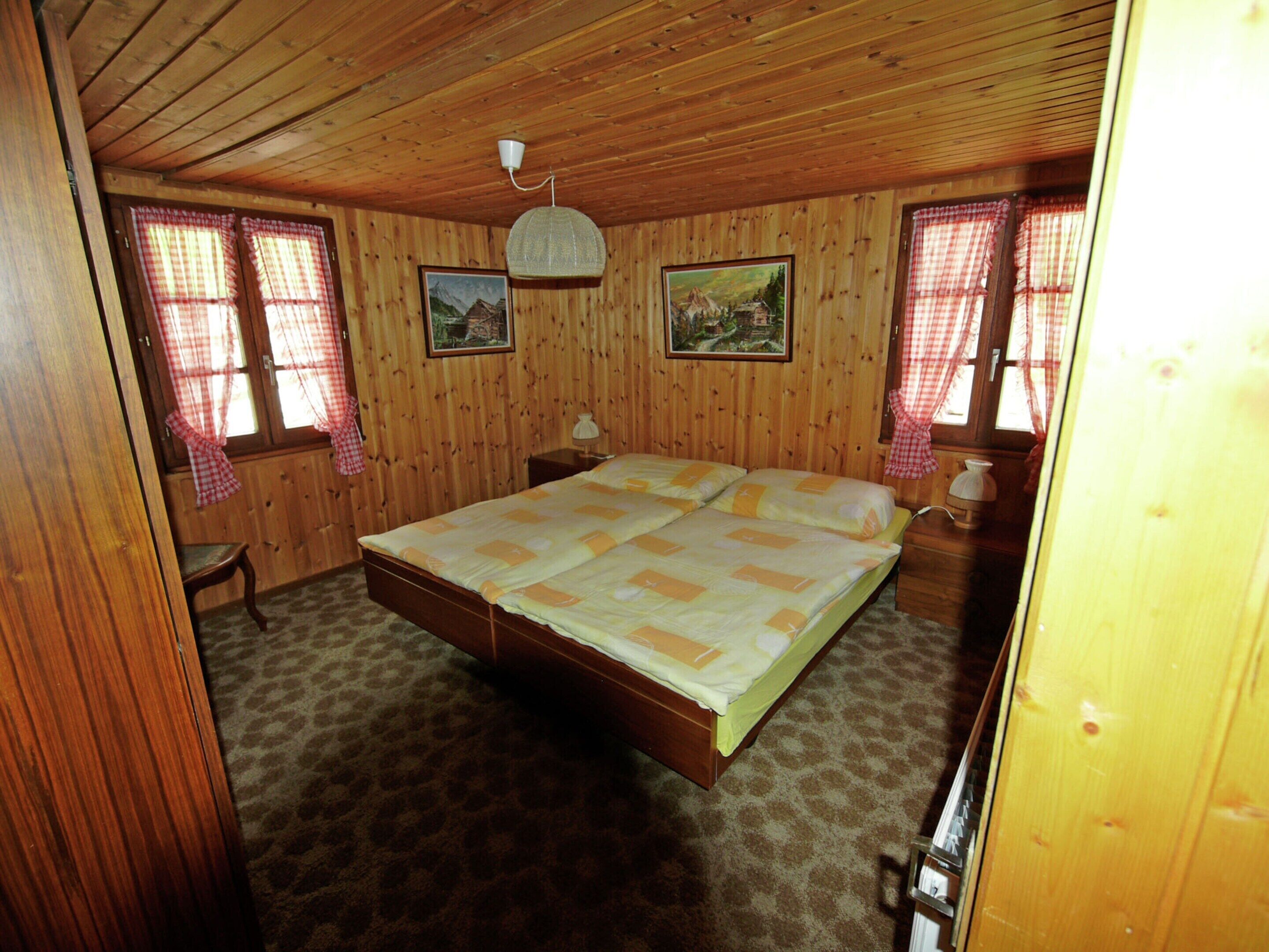 A wooden-paneled room with a large bed, two bedside tables, and two windows with red curtains.