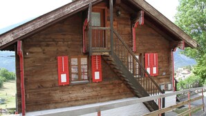 A wooden cabin with a wooden staircase leading to a balcony, red shutters, and a view of the mountains. - Boutique Apartment in Valais With Balcony (Guttet-Feschel)