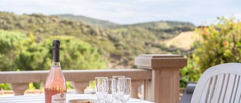 A table with a bottle, glasses, and a bowl on a terrace with a scenic view.
