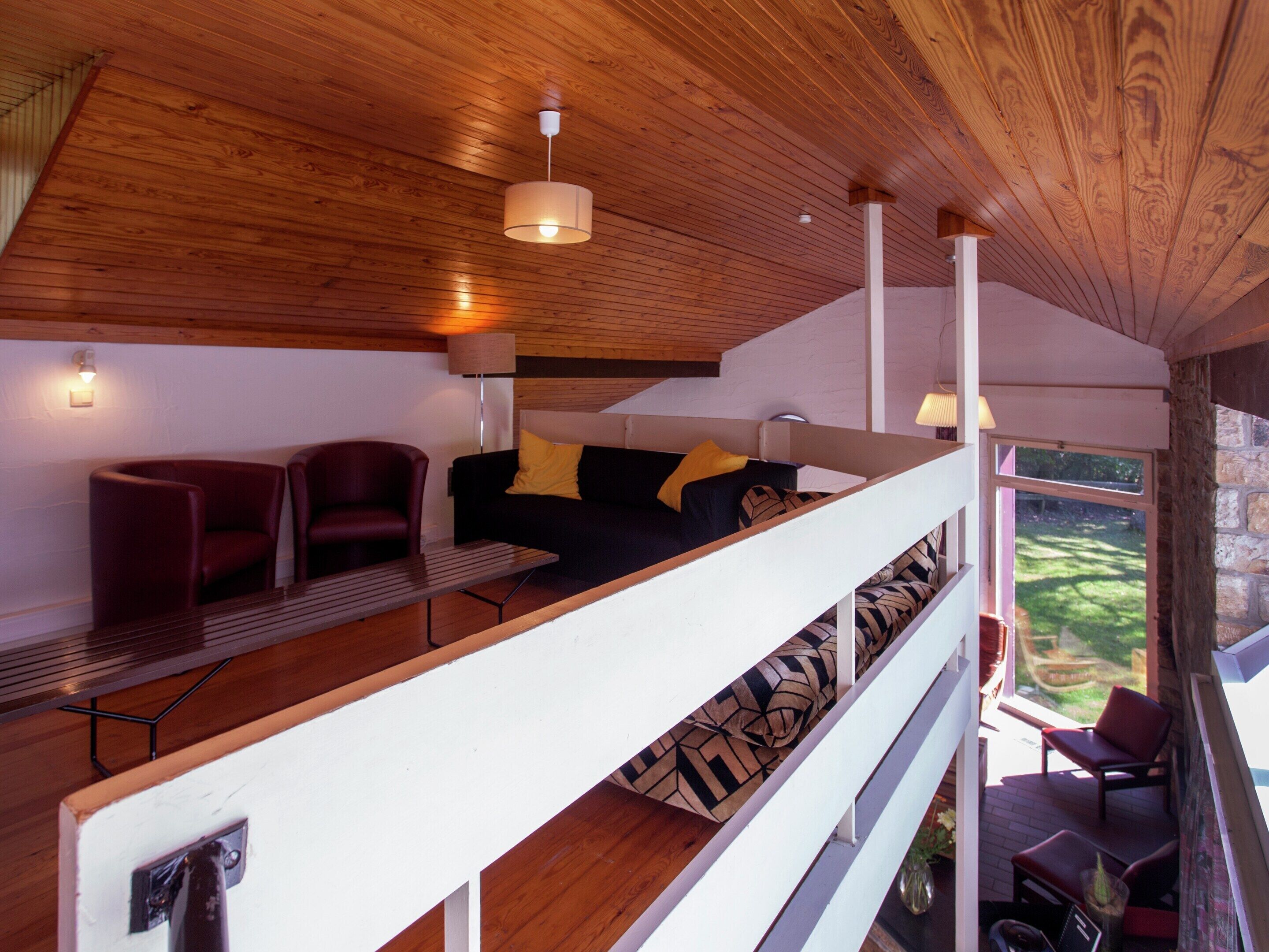 A modern interior with a wooden ceiling, a sofa with patterned cushions, and a view of a garden through a window.