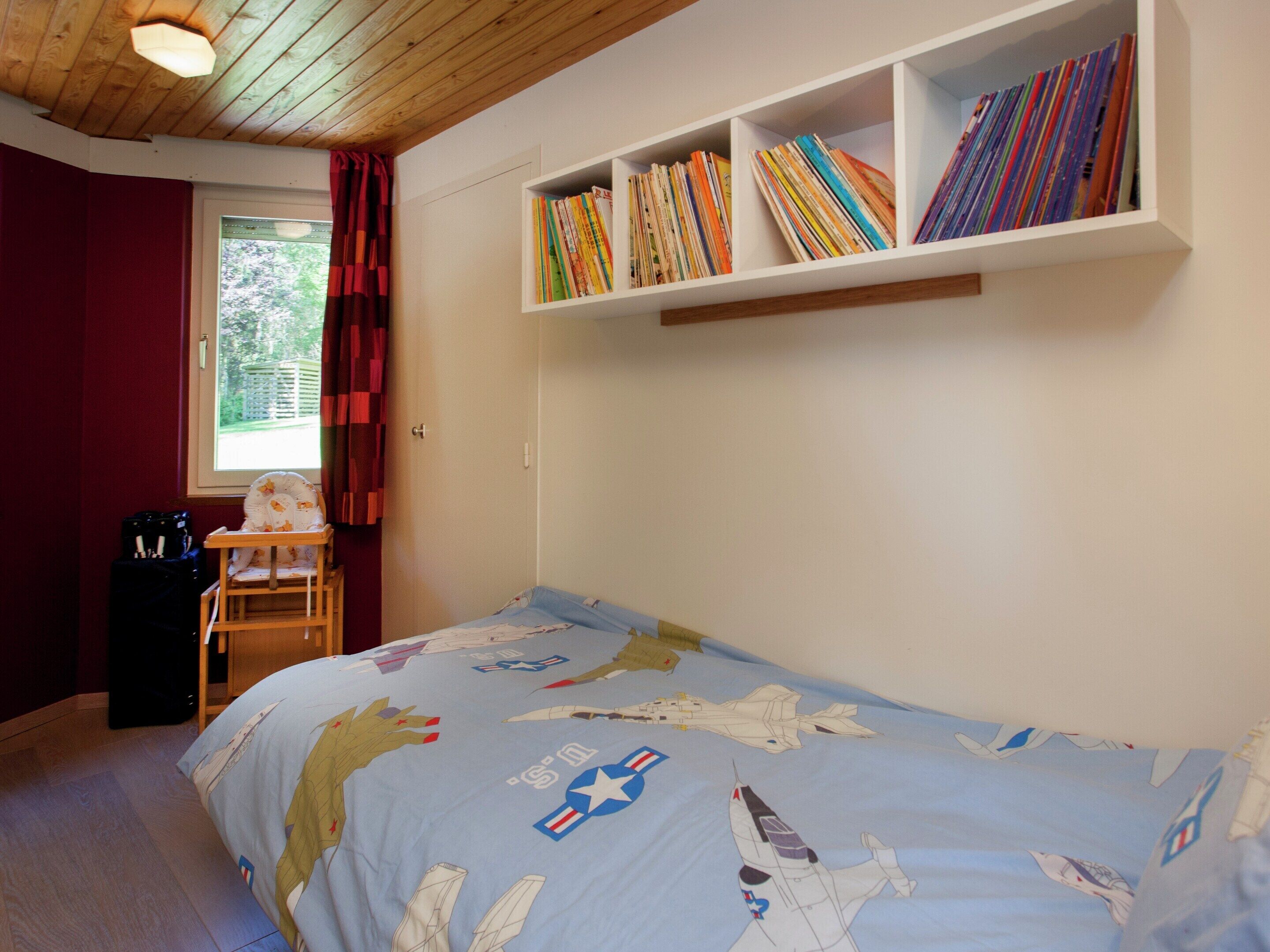 A bedroom with a bed featuring a space-themed bedspread, a bookshelf with colorful books, a window with red curtains, and a wooden ceiling.