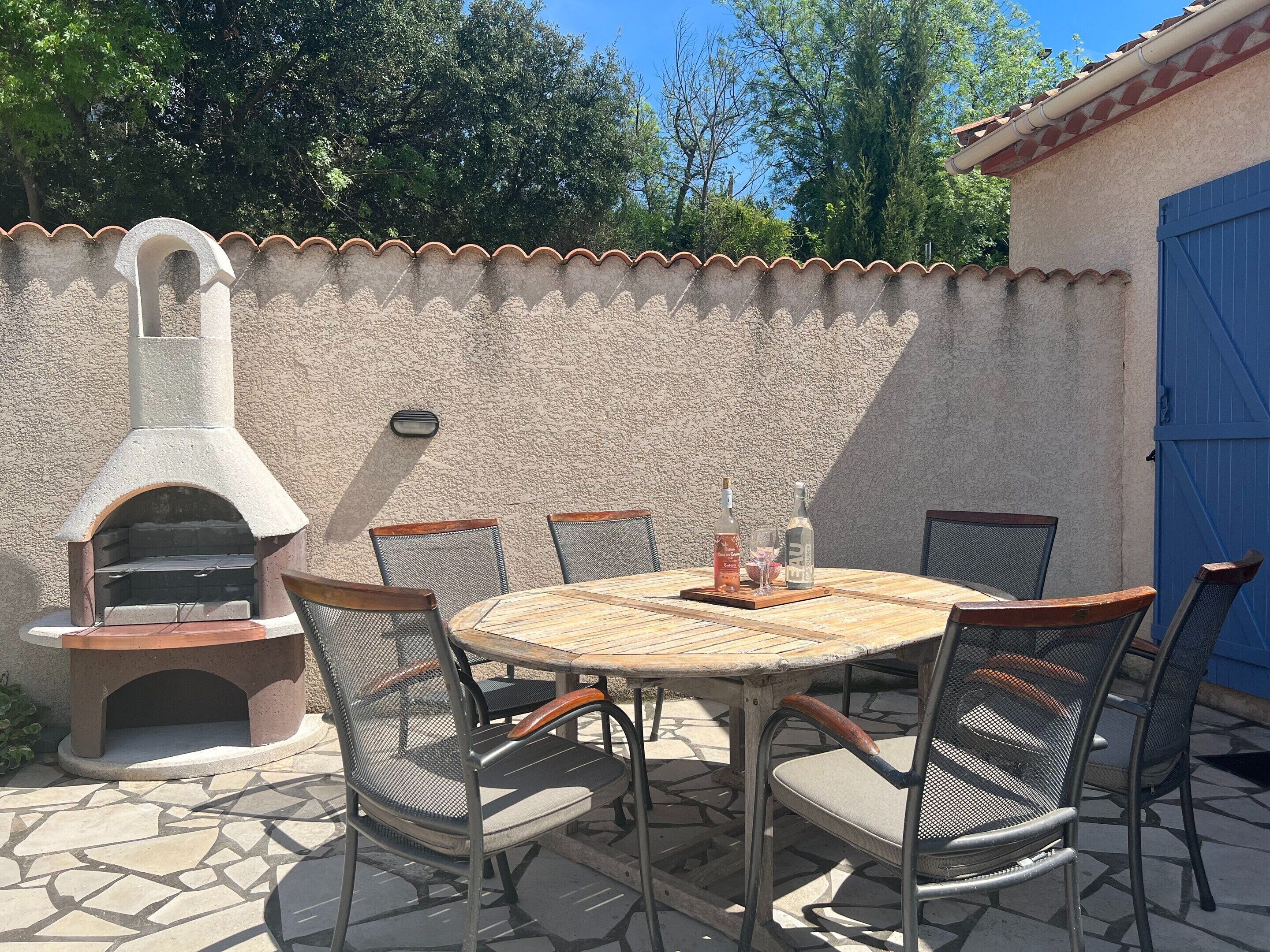 Une terrasse extérieure avec une table en pierre, six chaises en métal, un four à pizza et, au fond, une porte bleue.