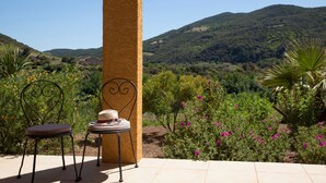 Eine Terrasse mit Stuhl und Hut, einer Säule sowie Blick auf eine gebirgige Landschaft.