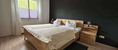 A bedroom with a wooden bed, a bedside table, a nightstand, a window with curtains, and a black wall.