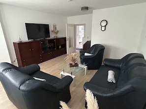 A living room with a TV, dark sofas, a glass coffee table, and a door leading to another room.