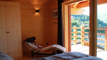 A wooden cabin room with a bed, a chair, a wooden door, and a large window with a view of a wooden deck and greenery.