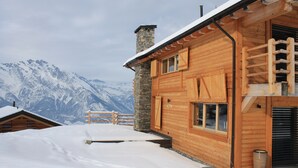 A wooden cabin with a stone chimney, wooden balcony, and a view of snow-covered mountains. - Chalet in La Tzoumaz Near Ski Lift (Riddes)