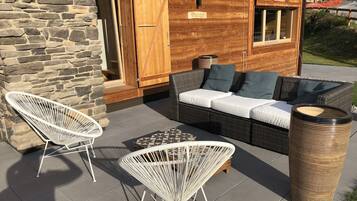 A wooden cabin with a stone wall, a wicker sofa with cushions, two white wire chairs, and a woven trash bin.