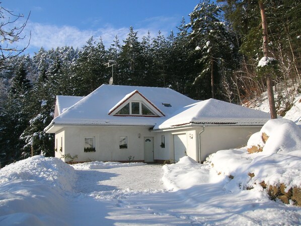 Ett snötäckt hus med ett triangulärt tak, omgivet av träd och ett snöigt landskap.