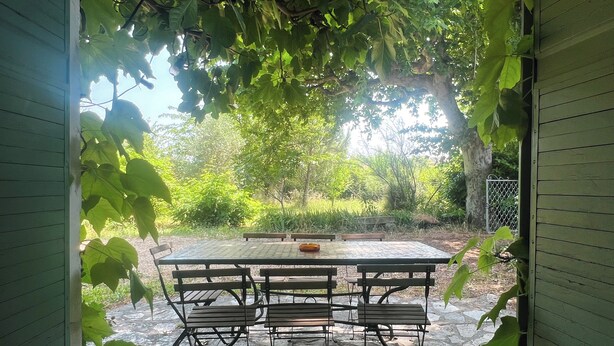 Une terrasse extérieure avec une table en pierre et des chaises en métal, entourée d’une végétation luxuriante et d’une clôture en bois.