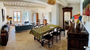 Un grand espace à manger avec une longue table et des chaises, un coin cuisine équipé d’une cuisinière et de placards, et un salon avec un canapé et une fenêtre.